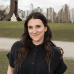 Kaela Blahey wearing a black button up shirt outside in front of green grass with a path halfway through. There are buildings in the background and greyish blue skies.