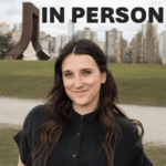Kaela Blahey wearing a black button up shirt outside in front of green grass with a path halfway through. There are buildings in the background and greyish blue skies. There is also the words "in person" in the sky because this is an in person workshop.